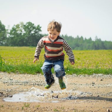 A cheerful child in a striped sweater jumps into a puddle on a sunny day in Czechia.