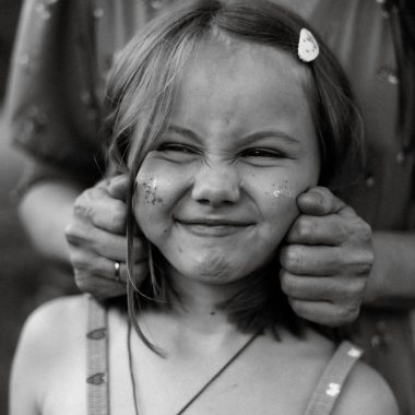 Charming black and white photo of a young girl making a playful face, with an adult's hands gently holding her cheeks.