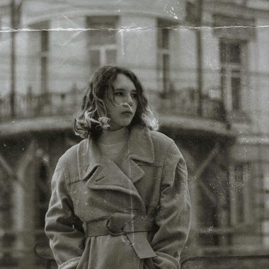 Artistic black and white portrait of a woman in a vintage coat outdoors against a historic building.